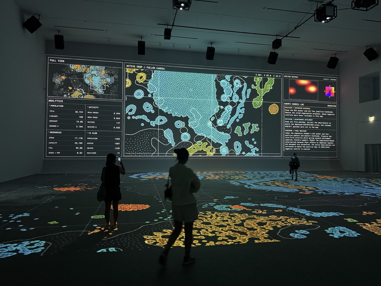 Visitors inside a dark gallery with colorful simulation patterns projected across a wall and floor.
